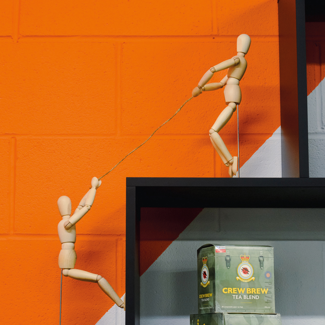 Two bendable wooden figures on the shelving unit - appearing as thought one is pulling the other up a step. Also in view, the Lincolnshire Tea Crew Brew packaging designed by our team